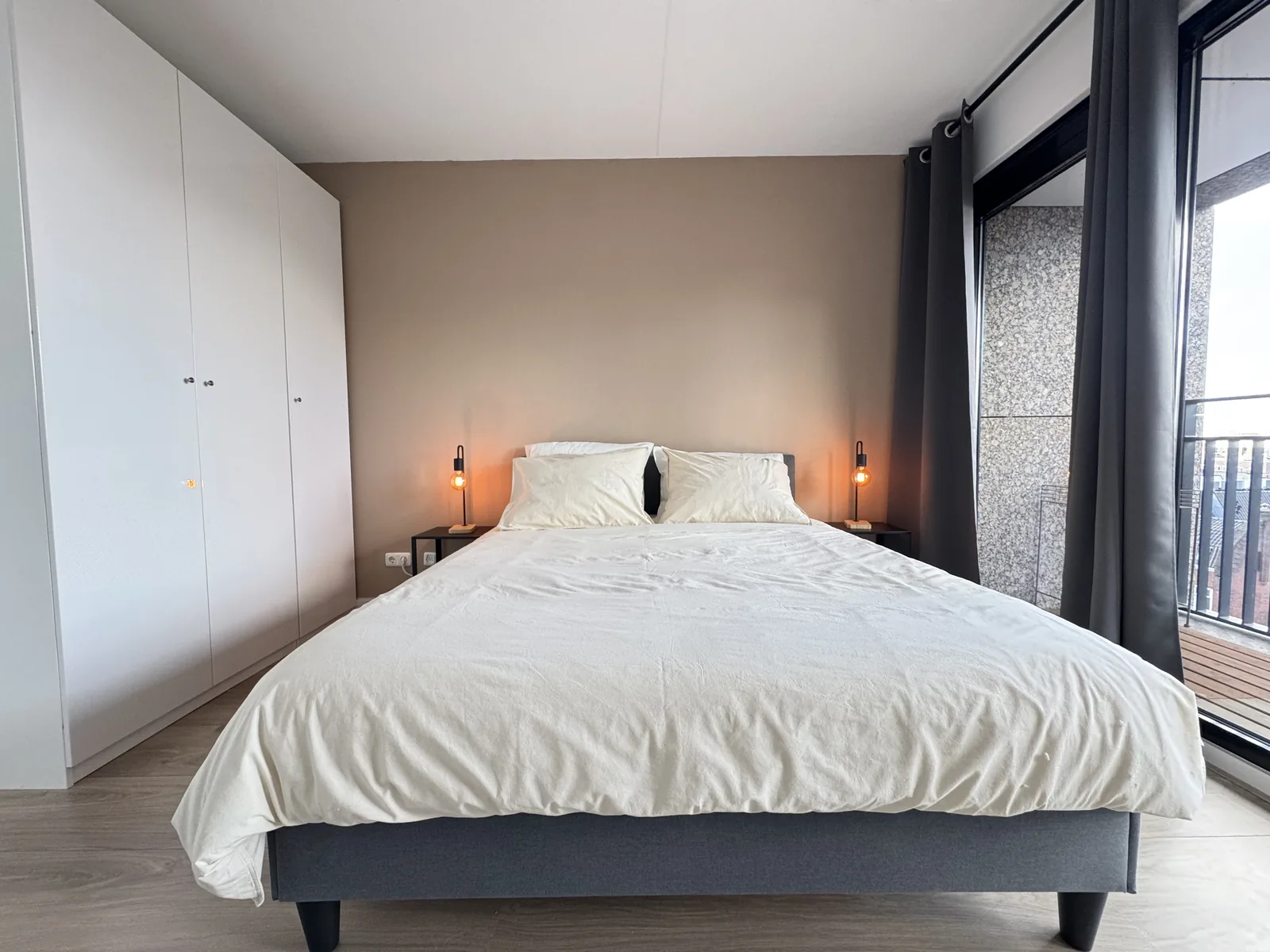 Master bedroom with large white wardrobe, double bed, and modern industrial bedside lamps in furnished apartment